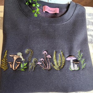May include: A dark gray sweatshirt with an embroidered design of mushrooms and ferns. The text "Cutemoon" is embroidered on the top left corner of the sweatshirt.