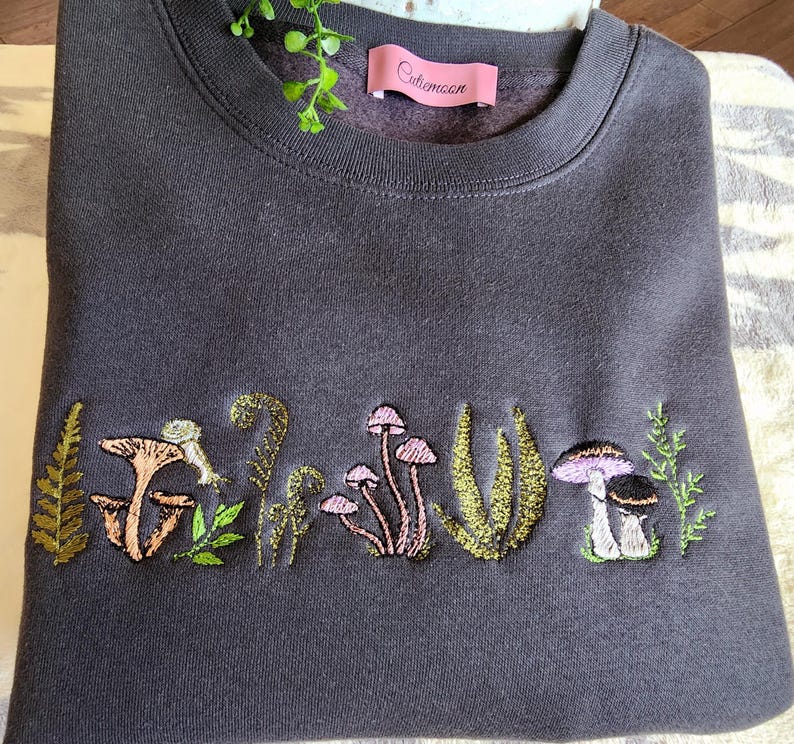 May include: A dark gray sweatshirt with an embroidered design of mushrooms and ferns. The text "Cutemoon" is embroidered on the top left corner of the sweatshirt.