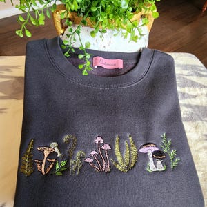 May include: A dark gray sweatshirt with an embroidered design of mushrooms and ferns. The sweatshirt has a pink label with the text "Oldmoon" on the front.