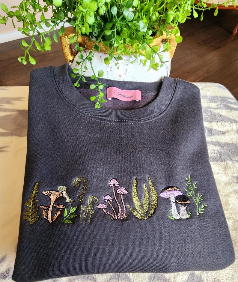 May include: A dark gray sweatshirt with an embroidered design of mushrooms and ferns. The sweatshirt has a pink label with the text "Oldmoon" on the front.
