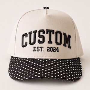 May include: A cream-colored baseball cap featuring the black embroidered text CUSTOM EST. 2024 on the front panel. The cap has a black brim decorated with a white polka dot pattern and a small black and white polka dot button on the top crown.