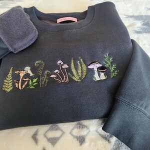 May include: A dark gray sweatshirt with a mushroom and fern embroidery design. The embroidery is in shades of green, pink, and brown.