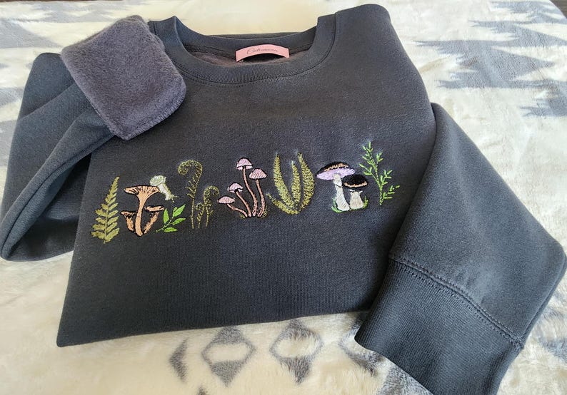 May include: A dark gray sweatshirt with a mushroom and fern embroidery design. The embroidery is in shades of green, pink, and brown.