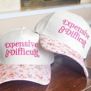 Peut inclure: Deux casquettes de baseball blanches avec le texte brodé "Expensive & Difficult" en rose. La visière de chaque casquette présente un motif floral rose. Les casquettes sont posées sur une surface en bois.