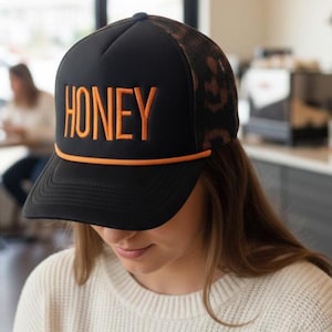 May include: Black trucker hat with the word "HONEY" embroidered in orange block letters. The hat features a black front panel, a mesh back with a leopard print design, and an orange rope detail along the brim.