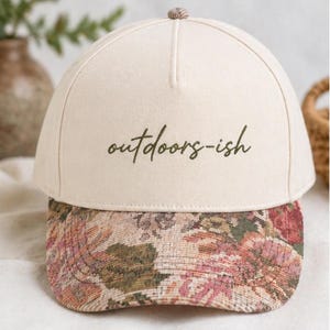 May include: A cream-coloured baseball cap with the text "outdoors-ish" in a dark green script. The brim has a floral tapestry design in pink, green, and brown. The top button is patterned fabric.