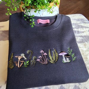 May include: A dark gray sweatshirt with an embroidered design of mushrooms and ferns. The text "Coleman" is on the tag.