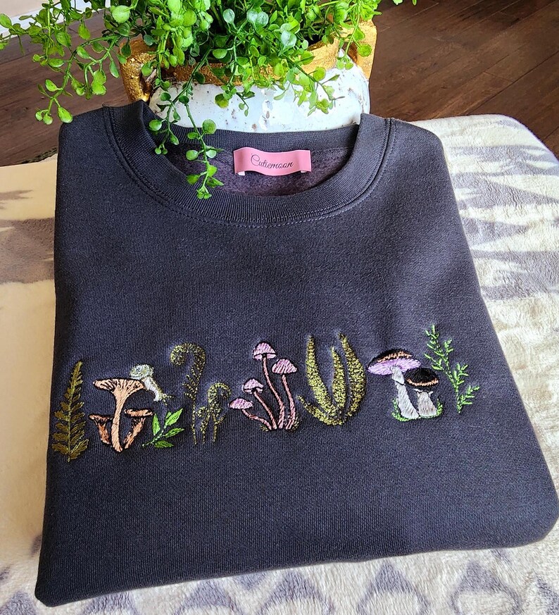 May include: A dark gray sweatshirt with an embroidered design of mushrooms and ferns. The text "Coleman" is on the tag.