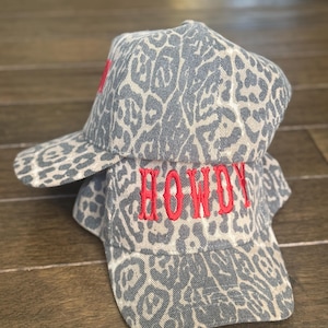 May include: Two baseball caps with a grey and white animal print pattern. The word "HOWDY" is embroidered in red on the front of the bottom cap. The caps are stacked on top of each other.