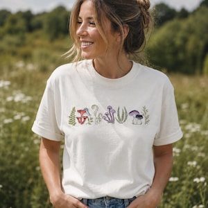 Può includere: T-shirt color crema con un design ricamato di vari funghi e piante nei toni del rosso, viola e verde. La maglietta è realizzata in un materiale morbido e viene indossata all'aperto.