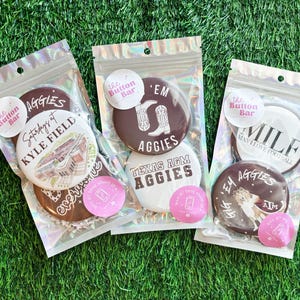 以下が含まれることがあります： 透明なプラスチックパッケージに入ったいくつかのボタンセットが、緑色の表面に表示されています。ボタンには、「Aggies」、「Kyle Field」、「MILF」などのテキストとグラフィックが使用されています。各パッケージには、「The Button Bar」と印刷された小さな丸いステッカーが付いています。