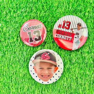 May include: Three baseball-themed buttons on green turf. One button is red with "Cornett 13" in white. Another features a photo of a boy with a baseball bat and the text "#13 Tildan Cornett". The third button has a boy's face and a baseball cap.