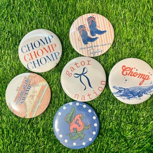 Puede incluir: Una colección de botones redondos con varios diseños y texto, incluyendo "CHOMP", un caimán y botas de vaquero. Los botones son blancos, azules, rojos y naranjas, y están colocados sobre hierba verde.