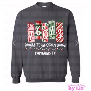 May include: Dark grey sweatshirt featuring a Christmas design. The design includes the numbers 76073, the words "Small Town Christmas" and "Paradise, TX". The design incorporates Christmas patterns and colours, with the text in white.
