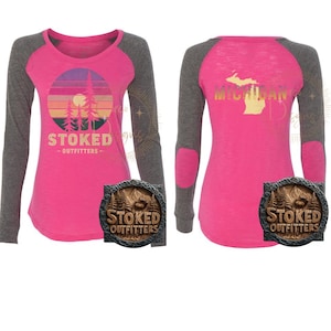 Puede incluir: Una camiseta rosa de manga larga con mangas raglán grises. La parte delantera presenta un gráfico de puesta de sol con árboles y el texto "STOKED OUTFITTERS". La parte trasera muestra un contorno dorado de Michigan y la palabra "MICHIGAN". Se muestra un letrero de madera con el mismo logotipo.