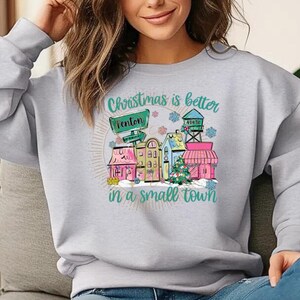 Pode incluir: Camisola cinza claro com um gráfico colorido que diz "Christmas is better in a small town". O gráfico apresenta uma cena da cidade com casas, uma árvore de Natal e um sinal que diz "Fenton".