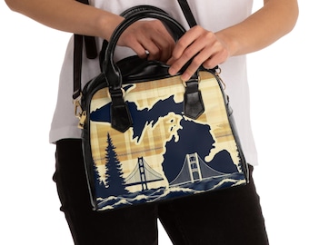 May include: A black leather handbag with a gold zipper and a printed design featuring a map of Michigan with a bridge and trees. The bag has a shoulder strap and a top handle.