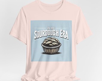 Puede incluir: Una camiseta rosa con un gráfico cuadrado azul que dice "In My Sourdough Era" y presenta un dibujo de una hogaza de pan de masa madre en una cesta.