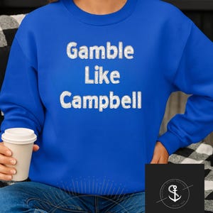 Peut inclure: Un sweat-shirt bleu roi avec l'inscription blanche "Gamble Like Campbell" en lettres stylisées. Le sweat-shirt est porté avec un jean bleu. Un petit logo est visible dans le coin inférieur droit.