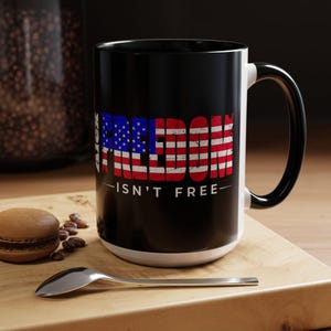 May include: Black ceramic mug with a white interior and handle, featuring the word "FREEDOM" in a distressed American flag design. The text "ISN'T FREE" is below the flag graphic. A spoon and a pastry are on a wooden surface.