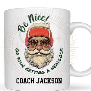 Puede incluir: Taza de cerámica blanca con una imagen de dibujos animados de Papá Noel con guantes de boxeo y gafas de sol. El texto "Be Nice! Or Your Getting A Headlock Coach Jackson" está impreso en la taza.