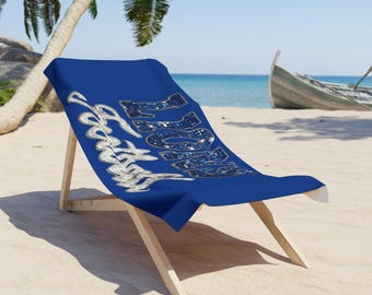 May include: A blue beach chair with a blue towel draped over it. The towel has the words "Beach Vibes" in a silver, cursive font. The chair is made of light-colored wood and is set on a sandy beach with a boat in the background.