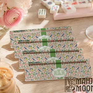 Op de afbeelding: Vier rechthoekige mahjong-tegelhouders met een bloemenpatroon van blauwe vogels, groene bladeren en roze bloemen. Elke houder heeft een centrale groene verticale streep met een wit geschulpt kader met het monogram MKM en de tekst MAHJ & MOON.