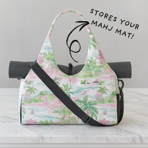 Può includere: Una borsa per tappetino da yoga con stampa tropicale in rosa, verde e blu. La borsa ha una tracolla e manici neri. Il testo "STORES YOUR MAHJ MAT!" è scritto sull'immagine.