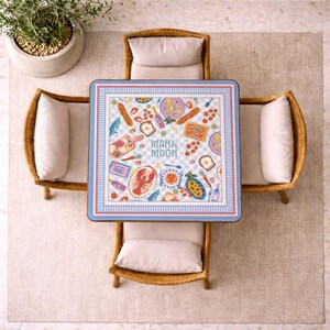 May include: A square table with a light blue and white checkered surface featuring colorful illustrations of food items, including a lobster, bread, and vegetables, with the text Mahj & Moon centered. The table is surrounded by four rattan chairs.
