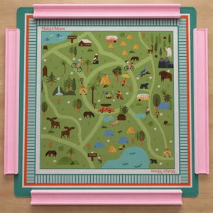 May include: A square Mahj&Moon game mat featuring a green illustrated map of a campground with tents, trees, wildlife, and vehicles. The mat is framed by a teal border with orange and white stripes and includes four pink plastic tile racks.