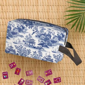 May include: A rectangular blue and white toiletry bag with a black handle. The bag features a scenic landscape design with pagodas and boats. Scattered around the bag are several purple mahjong tiles.