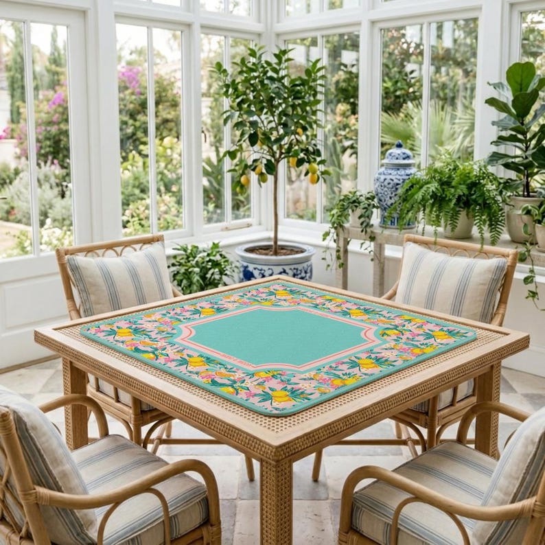 May include: A square table mat featuring a solid teal center bordered by a vibrant pattern of yellow lemons, pink flowers, and green leaves. The mat is placed on a light-colored wooden table surrounded by four matching rattan chairs with striped cushions.