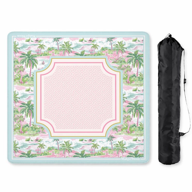 May include: A square mat featuring a tropical palm tree pattern in shades of green and pink, with a central light pink panel displaying a Greek key border and a light blue trim. The text Mahj & Moon is visible. A black carrying bag is included.