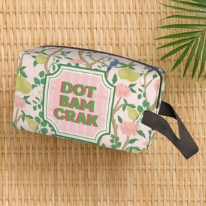 May include: A rectangular wash bag featuring a floral design with pears and pink flowers on a light pink backdrop. It has a black handle and the text "DOT BAM CRAK" within a pink and green frame.