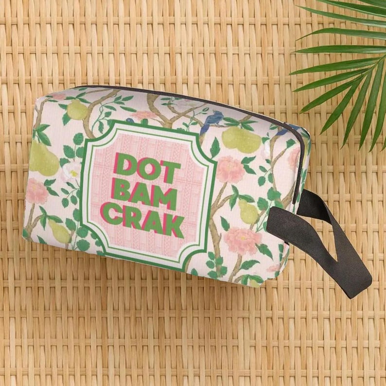 May include: A rectangular wash bag featuring a floral design with pears and pink flowers on a light pink backdrop. It has a black handle and the text "DOT BAM CRAK" within a pink and green frame.