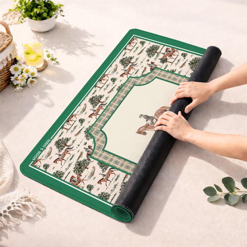 Puede incluir: Una alfombrilla rectangular con borde verde oscuro y centro color crema que presenta un patr&oacute;n ecuestre repetitivo de caballos y jinetes entre &aacute;rboles. La alfombrilla tiene un marco a cuadros y el texto "man & moon" impreso en el lado izquierdo.