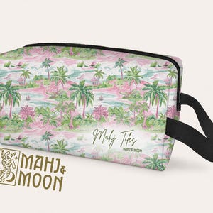 Op de afbeelding: Een rechthoekige toilettas met een tropische eilandprint met palmbomen, roze bloemen en turquoise water. De tas heeft een zwarte rits en handvat, met de tekst "Mahj Tiles" en "Mahj & Moon".