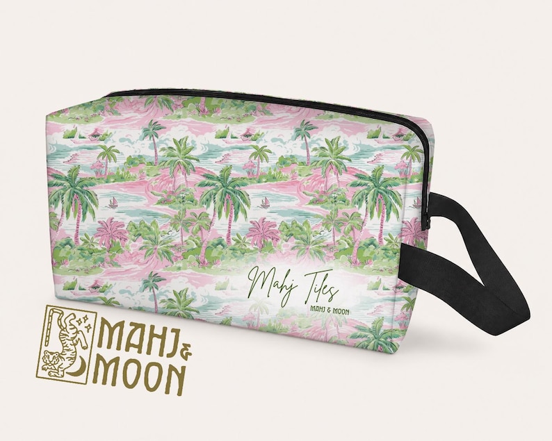 Op de afbeelding: Een rechthoekige toilettas met een tropische eilandprint met palmbomen, roze bloemen en turquoise water. De tas heeft een zwarte rits en handvat, met de tekst "Mahj Tiles" en "Mahj & Moon".
