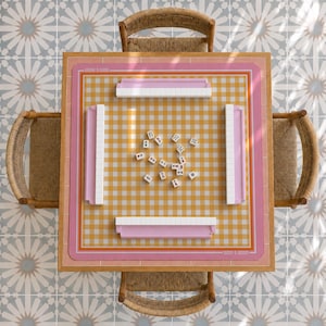 May include: A square wooden table with four woven chairs features a yellow and white gingham play mat with a pink border. Four pink tile racks are positioned on the mat, surrounding a central collection of white mahjong tiles with symbols.