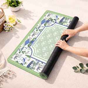 May include: A rectangular yoga mat with a sage green border and a white center featuring a blue and green pattern of elephants, trees, and architectural structures. The text "Mahji Moon" is printed on the mat. The underside is solid black.