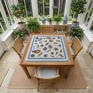 May include: A square table with a light-colored top featuring a colorful pattern of various food items, sea creatures, and drinks, surrounded by four light brown rattan chairs with blue and white striped cushions, set in a bright sunroom.