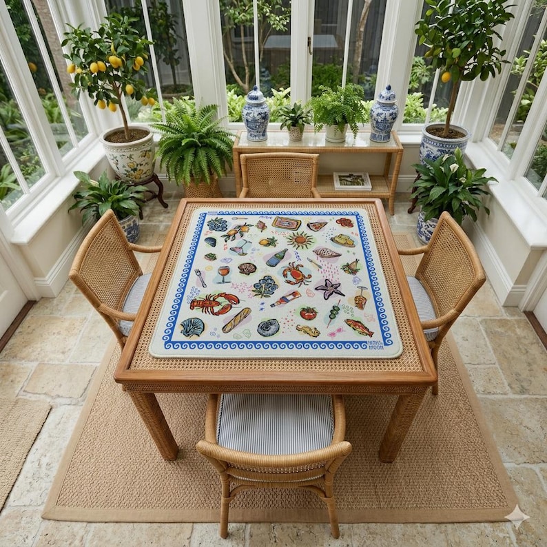 May include: A square table with a light-colored top featuring a colorful pattern of various food items, sea creatures, and drinks, surrounded by four light brown rattan chairs with blue and white striped cushions, set in a bright sunroom.