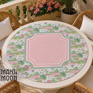 May include: A round tablecloth featuring a tropical landscape print with palm trees in shades of pink and green. The center displays a pink square with a white Greek key pattern, bordered by a light blue frame with the text Mahj & Moon.