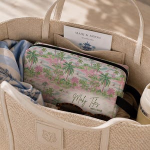 Op de afbeelding: Beige draagtas met cr&egrave;mekleurige handvatten, gevuld met een cosmetische tas met tropisch thema, een blauw-witte sjaal, zonnebril en een waterfles. De cosmetische tas heeft een roze en groen palmboomontwerp. Een kaart met "Mahi & Moon" is zichtbaar.