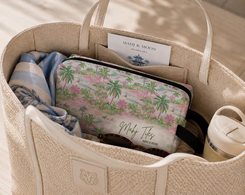 Op de afbeelding: Beige draagtas met cr&egrave;mekleurige handvatten, gevuld met een cosmetische tas met tropisch thema, een blauw-witte sjaal, zonnebril en een waterfles. De cosmetische tas heeft een roze en groen palmboomontwerp. Een kaart met "Mahi & Moon" is zichtbaar.