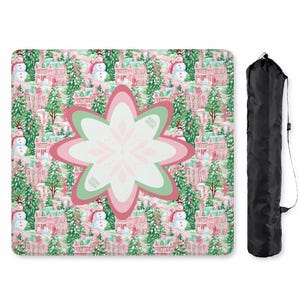 Op de afbeelding: Een vierkante deken met een herhalend winterpatroon van roze huizen, groene dennenbomen en sneeuwpoppen, met een grote, centraal geplaatste, meerlaagse bloemvormige afbeelding in roze, wit en groen. Een zwarte draagtas is inbegrepen.