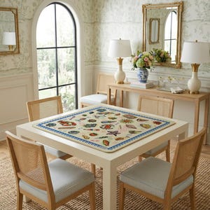 May include: A square cream-colored tablecloth featuring a colorful pattern of various seafood, including lobsters, crabs, and shellfish, bordered by a blue and white wave design, centered on a light-colored wooden dining table with four chairs.