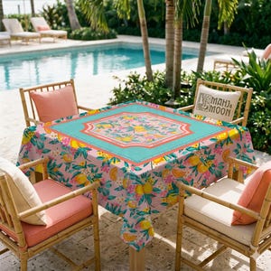 May include: A square pink tablecloth with a floral and citrus fruit pattern, centered with a turquoise border. The table is surrounded by bamboo chairs with coral cushions, set near a pool.