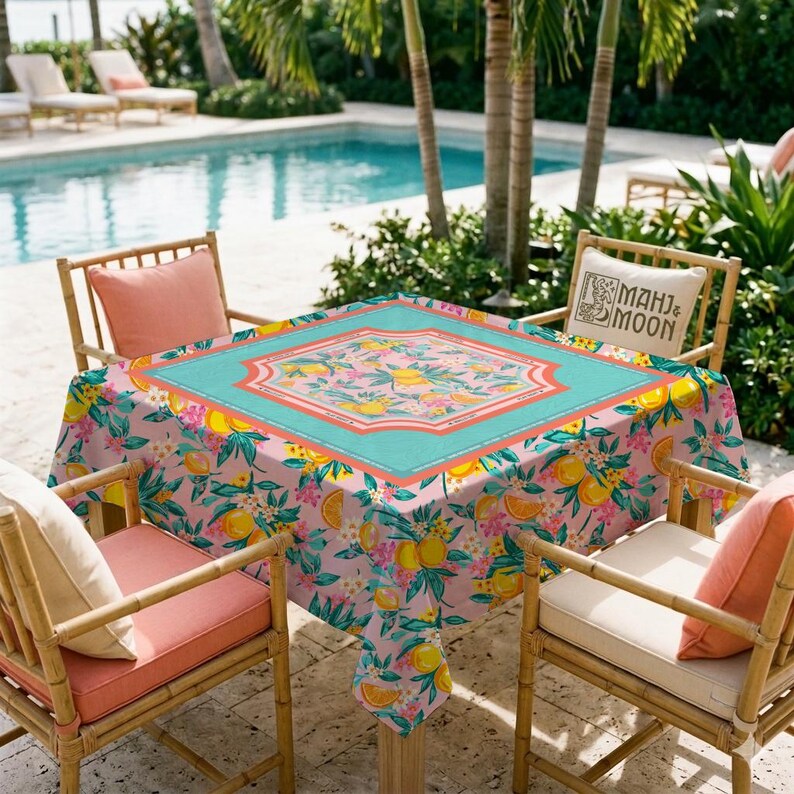 May include: A square pink tablecloth with a floral and citrus fruit pattern, centered with a turquoise border. The table is surrounded by bamboo chairs with coral cushions, set near a pool.