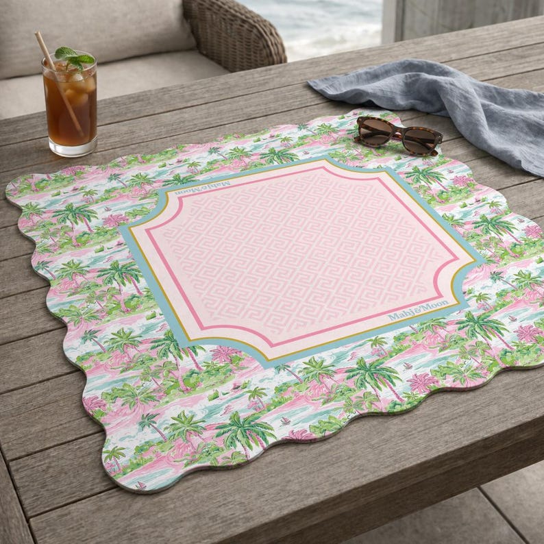 May include: A square placemat with scalloped edges featuring a tropical palm tree pattern in shades of pink and green. The centre has a light pink panel with a Greek key design, framed by a blue and gold border with the text Mahj & Moon.